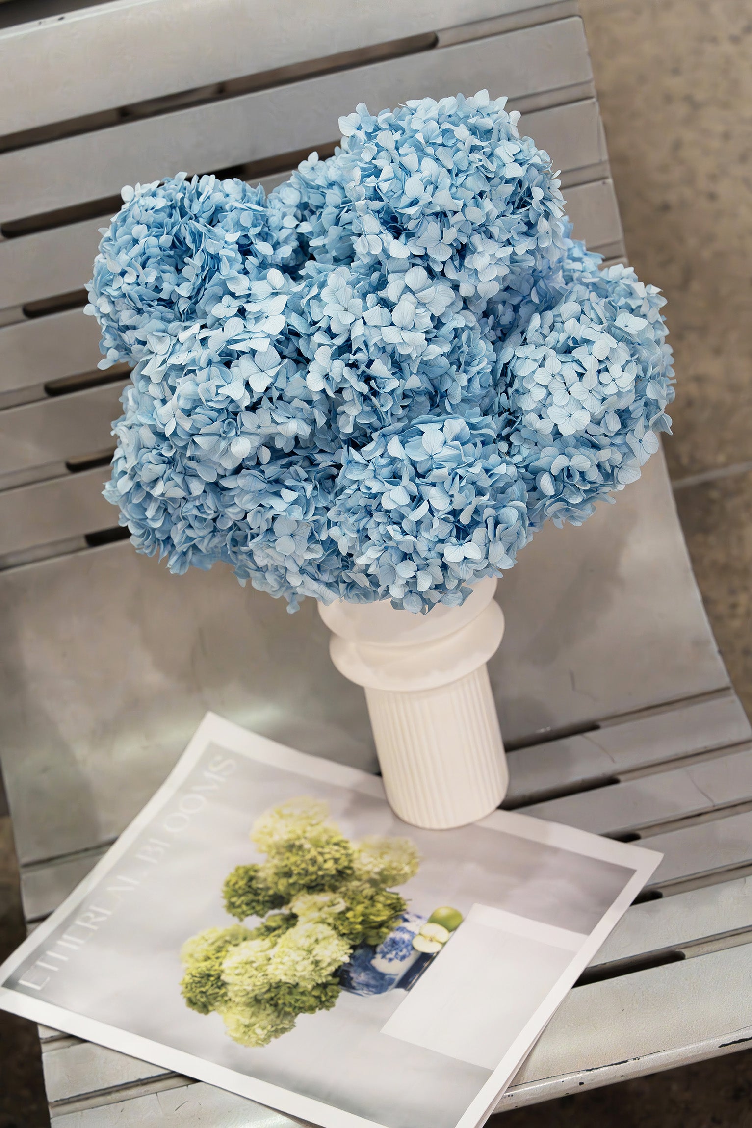 Bouquet of preserved blue hydrangea heads in a white vase with a newspaper underneath.